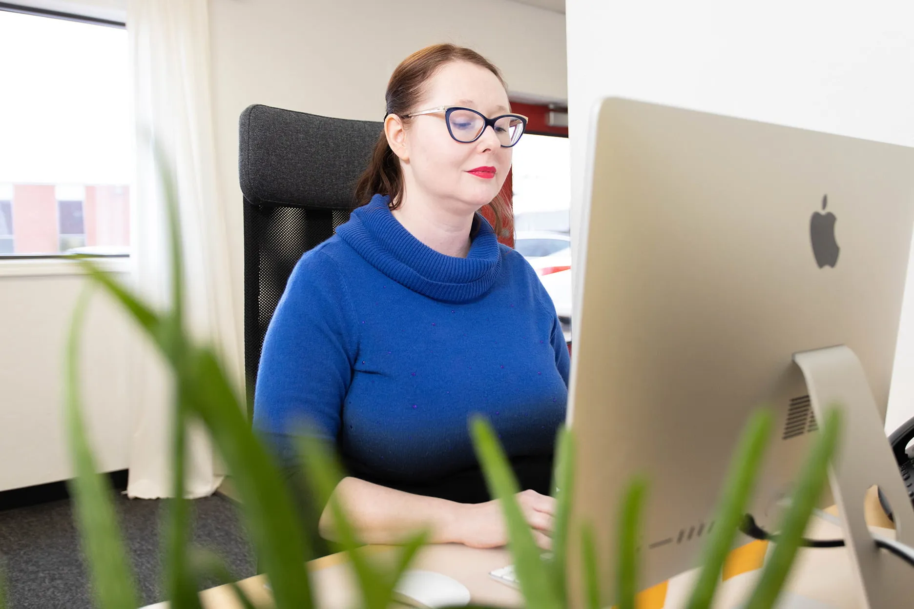 April types on her Mac while working on Oklahoma SEO services at her desk. She is wearing a blue sweater and red lipstick. 