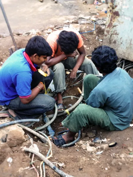 Three young men sit in a circle in the dirt and talk. 