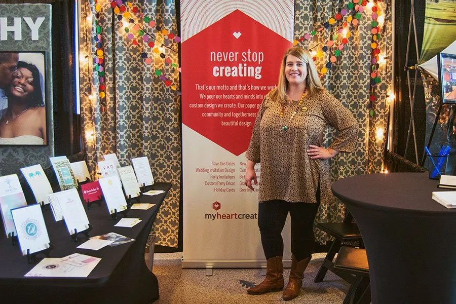 Amy smiles with her hand on her hip while standing in the myheartcreative booth at the Oklahoma Bridal Show. Behind her is a large retractable banner that says, "Never Stop Creating" at the top. On her right is a table with various print materials and wedding invitation samples. 
