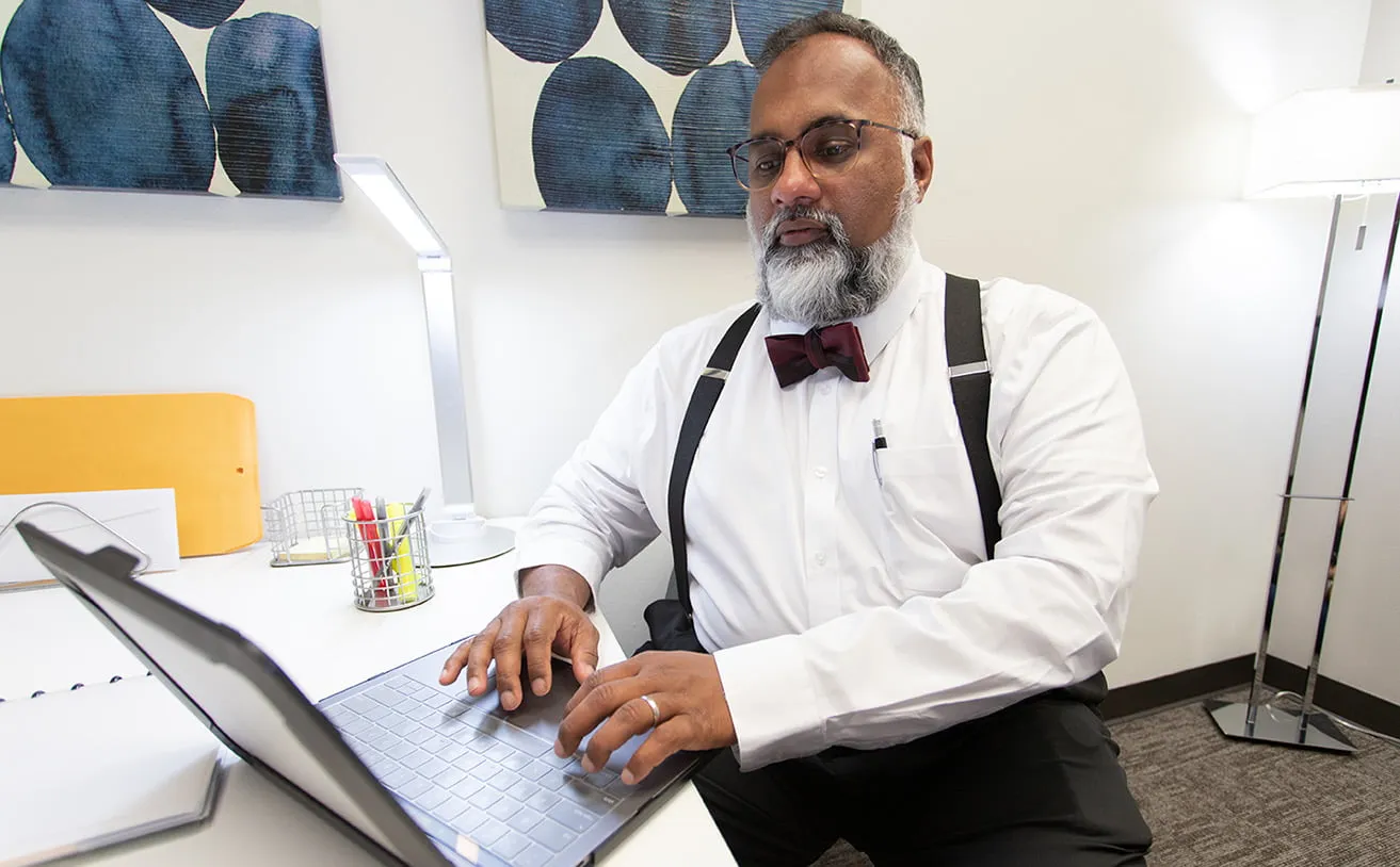 Sam, the Operations Director at myheartcreative, works on his laptop in his office at the studio. He is wearing suspenders and a maroon bow tie. 