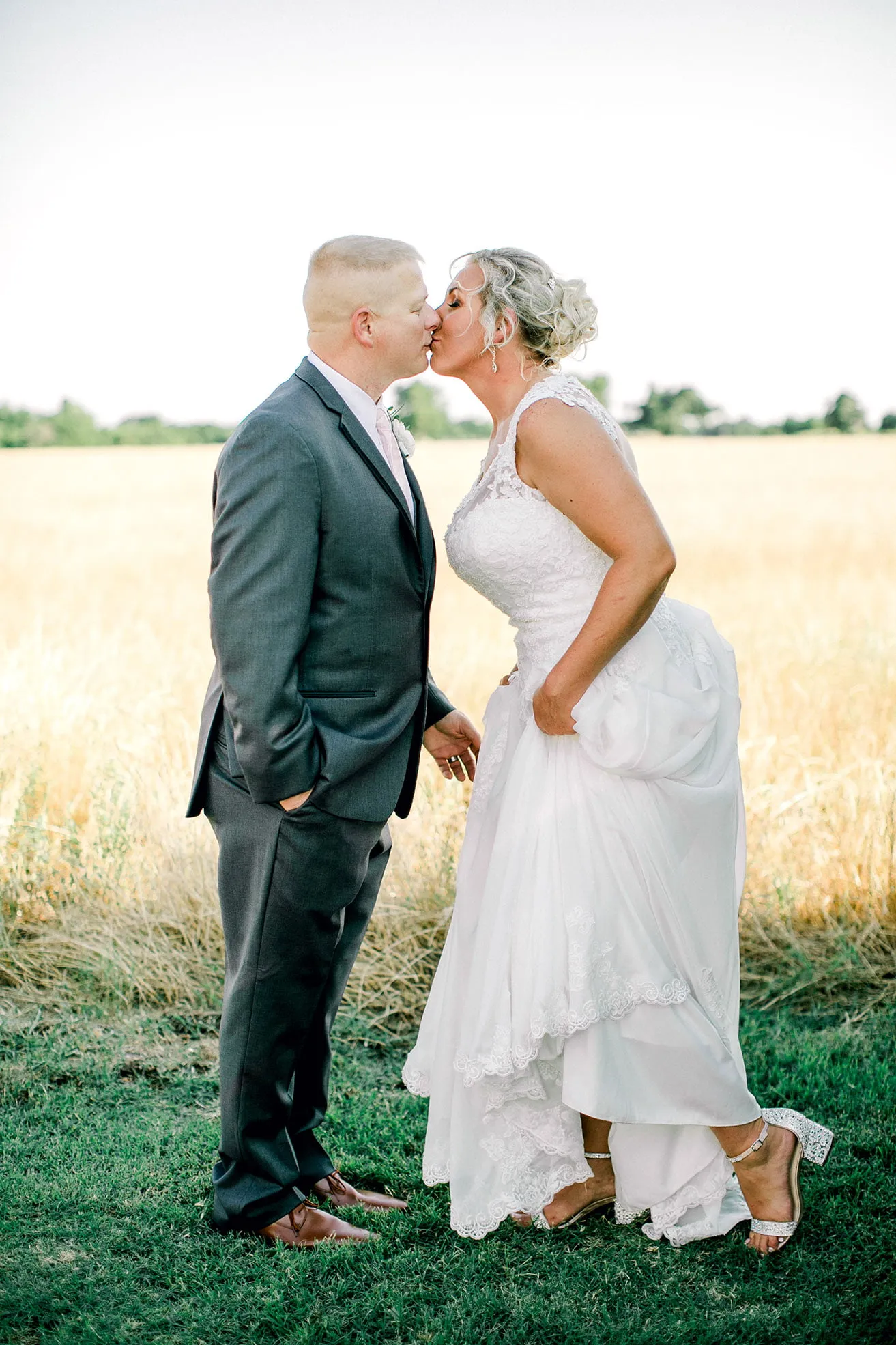 Jeff and Kasey The Palace Wedding - myheartcreative The bride leans forward and kisses the groom as they pose for photos next to a wheat field on their wedding day.