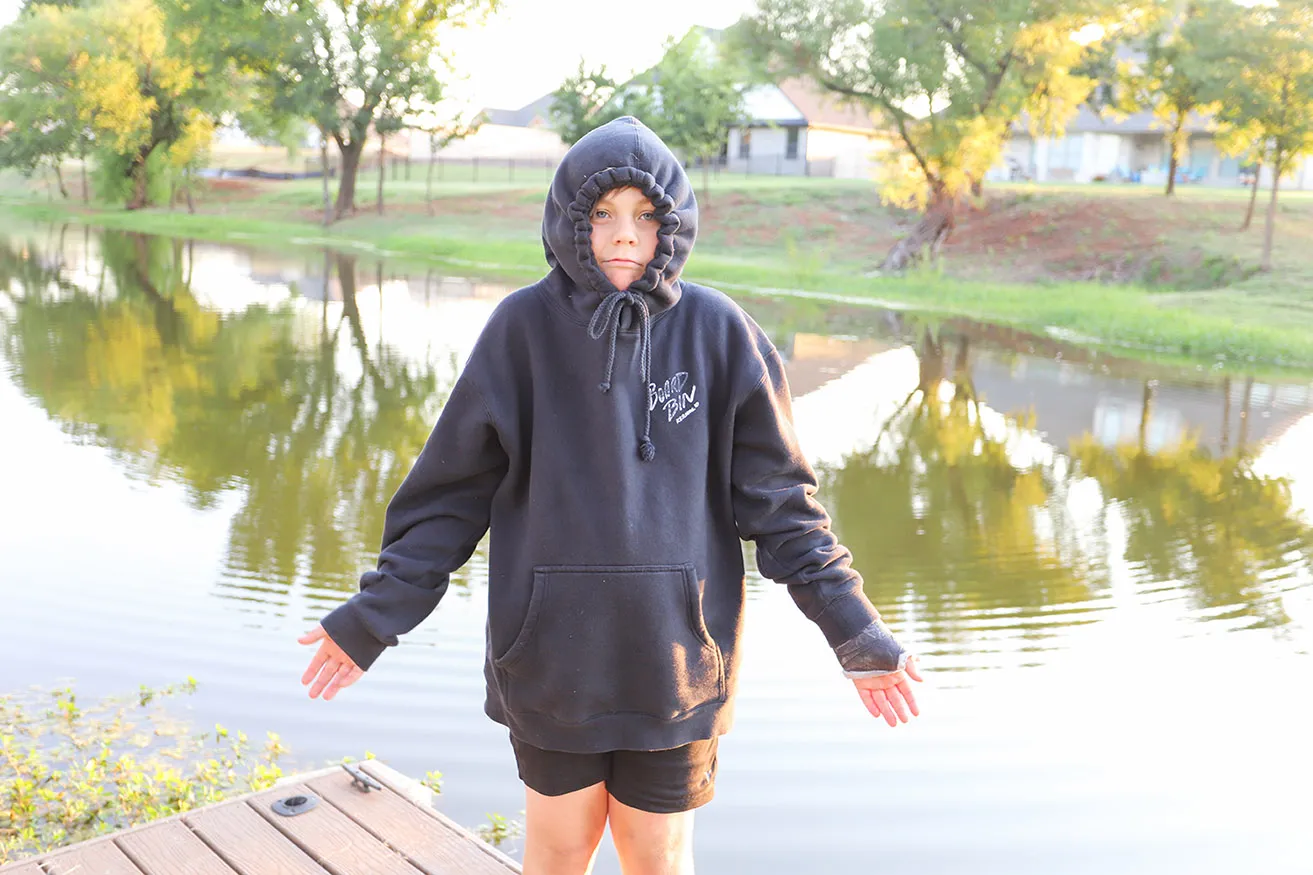 A young boy shrugs with his jacket hood over his head. He stands on a dock. 