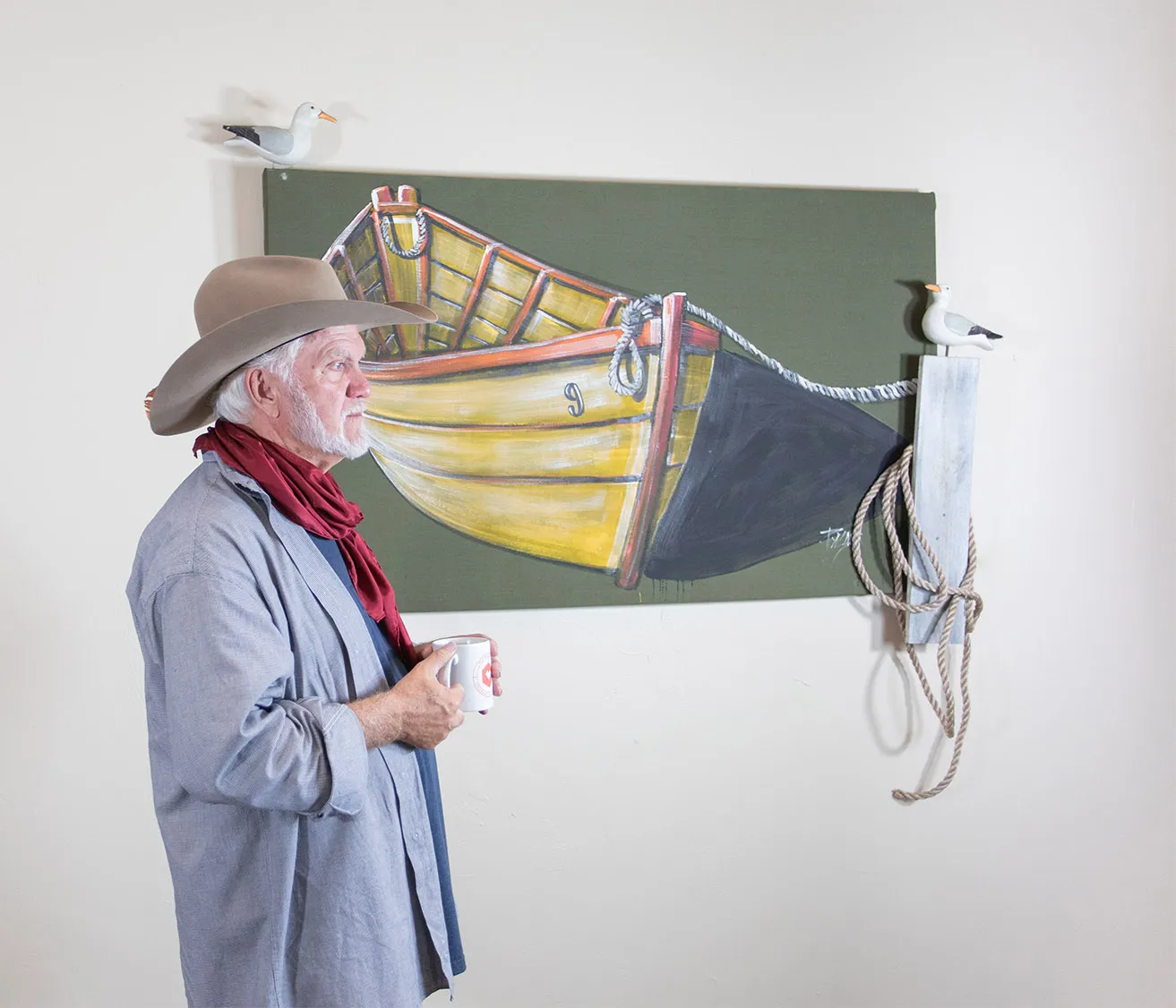 Dr. Palmer stands next to one of his paintings of a small boat while holding a cup of coffee. 