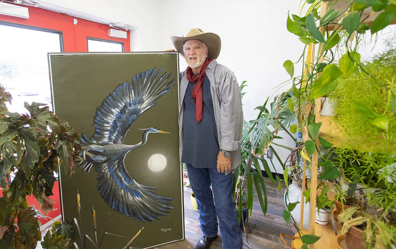 Artist Dr. Bob Palmer stands next to a large painting of a bird. He is surrounded by plants at the studio. 