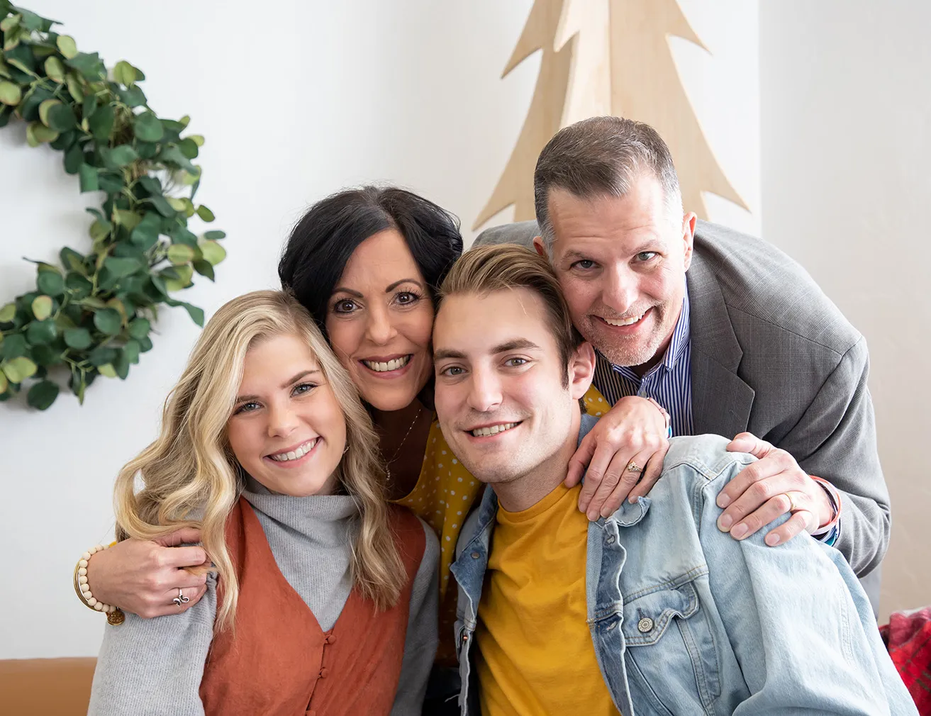 A family of four poses for a photo. The mother and father bend down and smile with the teenage daughter and son. They are wearing a harmonious color palette. 