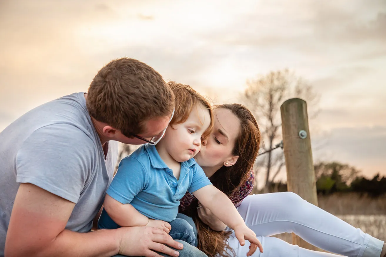 A mother and father both kiss each side of a toddler's face during an outdoor photoshoot. 