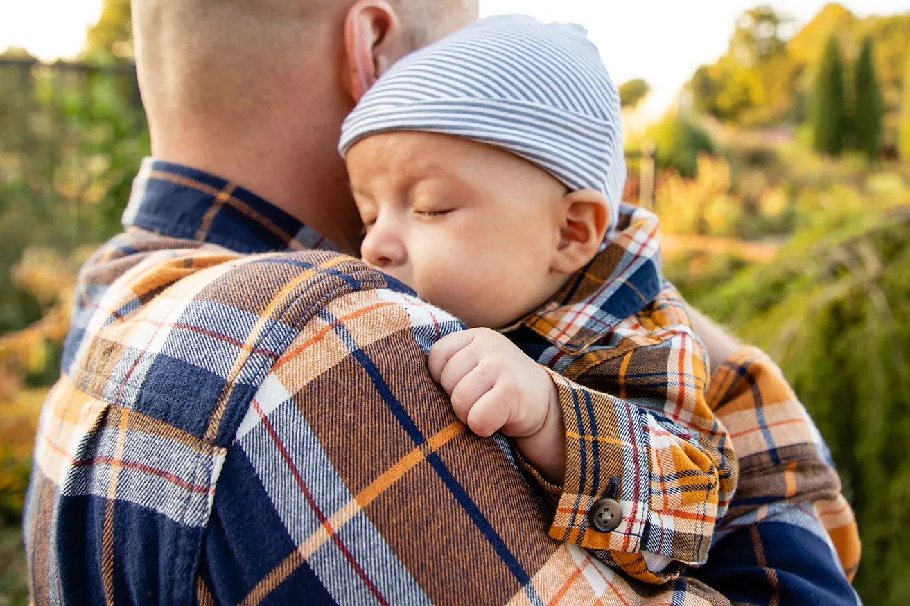 A newborn baby boy sleeps while resting on his dad's shoulder during a family photoshoot. 