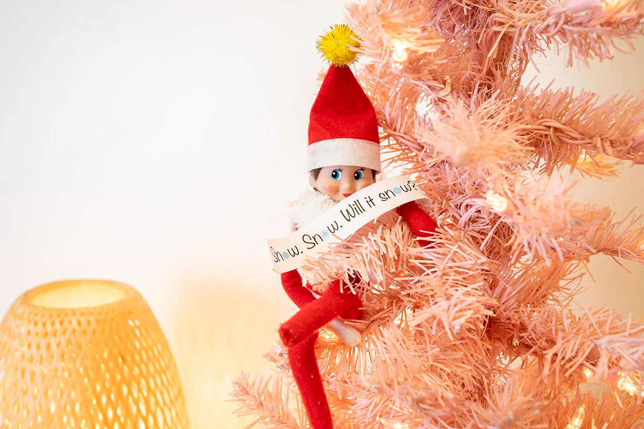 An Elf on the Shelf sitting in a pink tree with a printable around it that says, "Snow. Snow. Will it snow?"