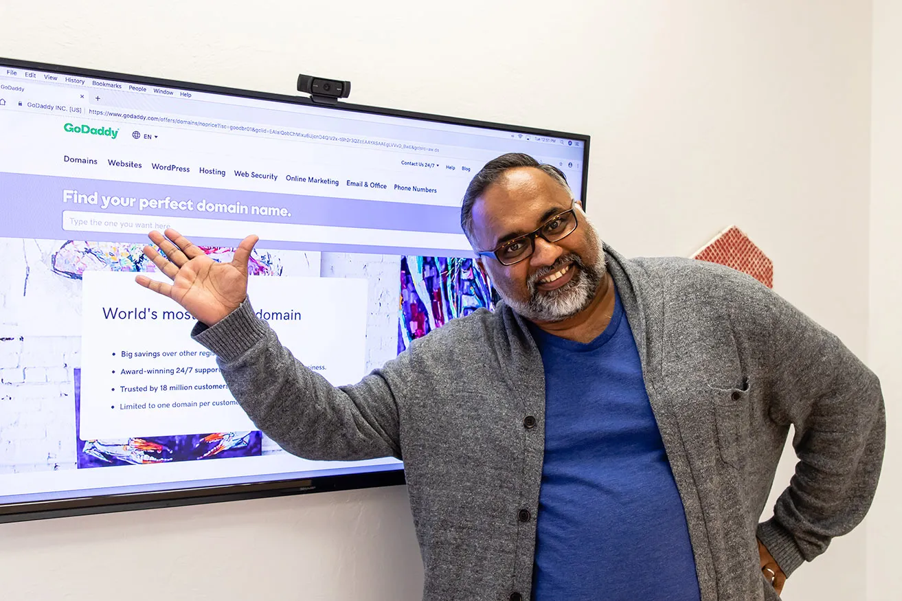 Sam, the Operations Director, points to the GoDaddy website on the conference room monitor. 