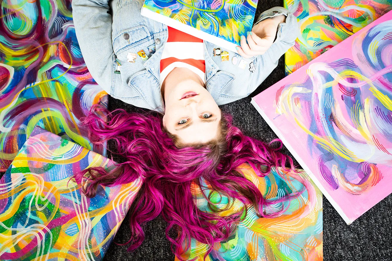Artist Chandler Domingos lays amongst several of her colorful abstract paintings. Her bright pink hair is spread out in all directions. 