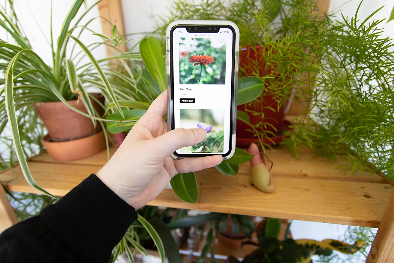 Kristen holding a smartphone while scrolling on SoulFly Doulas' online shopping cart. She is standing in front of a shelf with plants. 