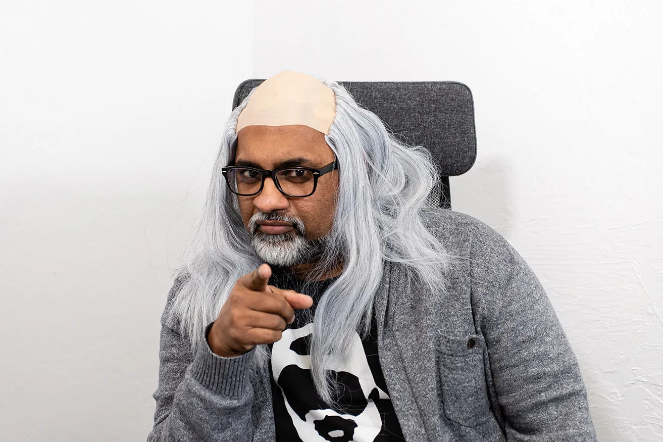 Sam is sitting at his desk in his office. He is pointing at the camera with a serious face while wearing a grey wig. 