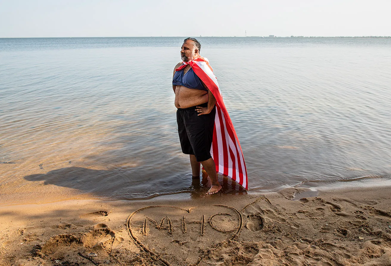 Sam stands on the edge of a lake with his t-shirt rolled up to mimic a bathing suit top. He wears the American flag as a cape and has written "MHC" in a heart in the sand. 