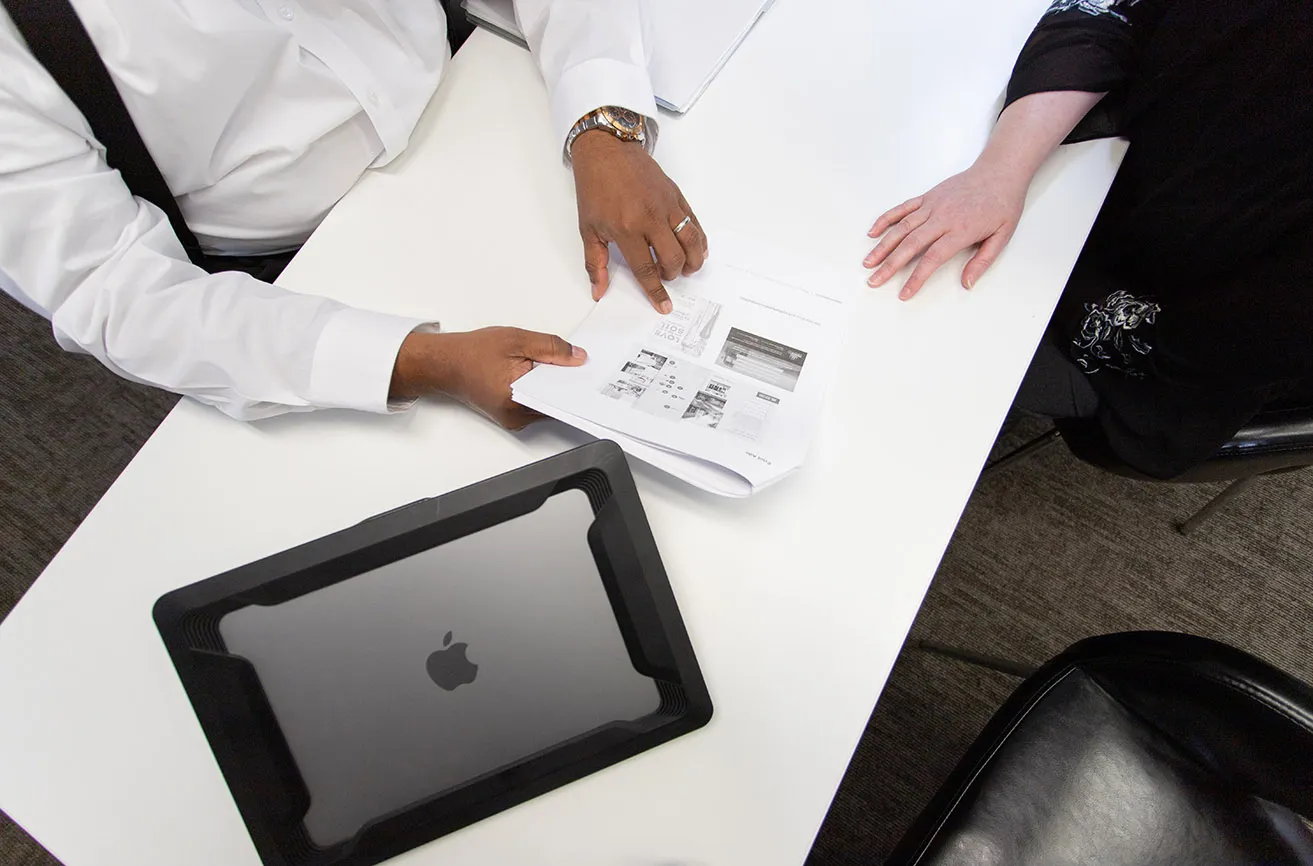 Is Digital Marketing OKC Worth It in 2021? - myheartcreative April and Sam discussing Oklahoma marketing results for a client at his desk in the studio. Sam's Apple Macbook rests next to the paper with results.