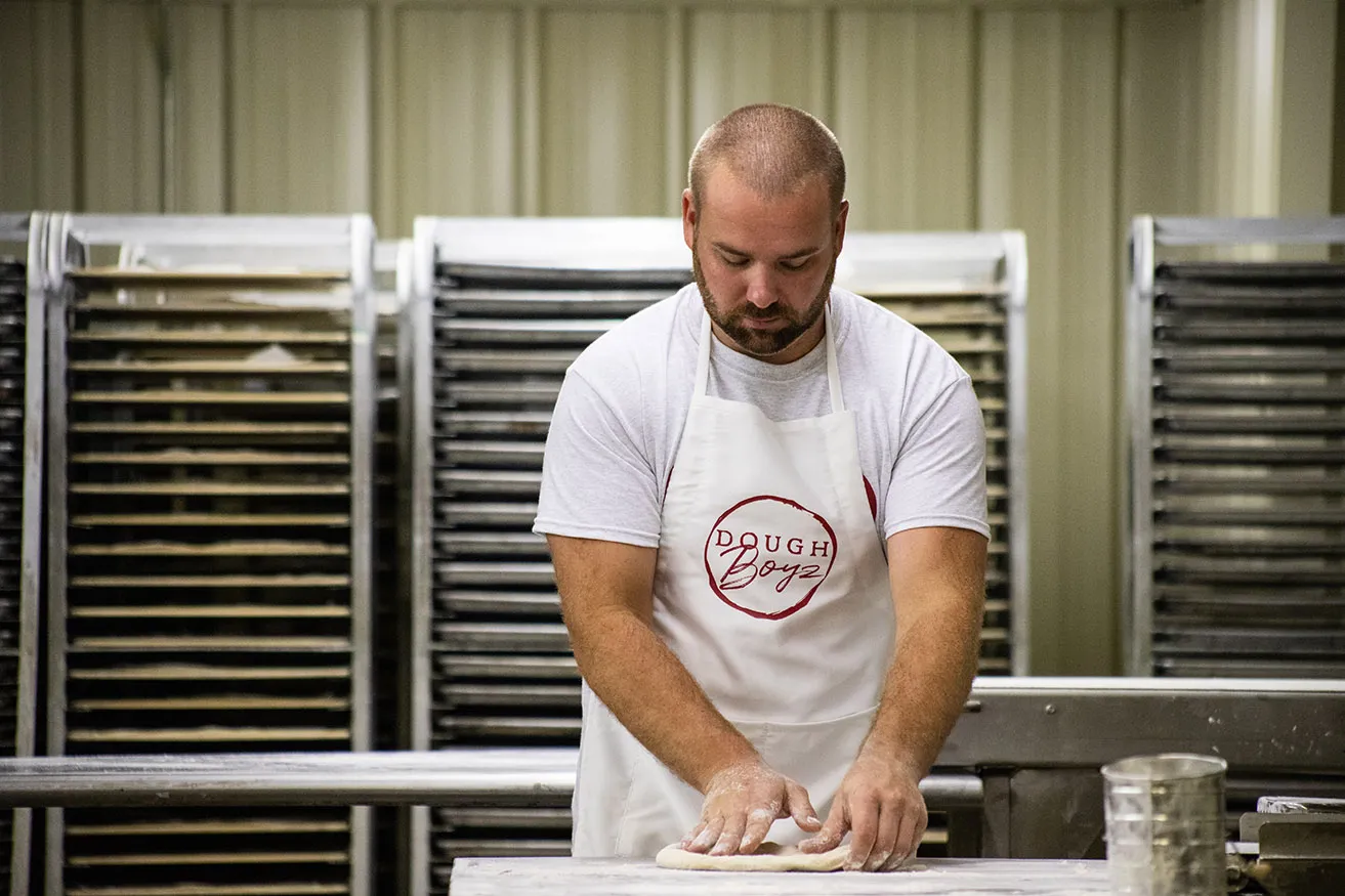 A pizza dough maker wearing a white apron with the DoughBoyz logo in red font on the front. 