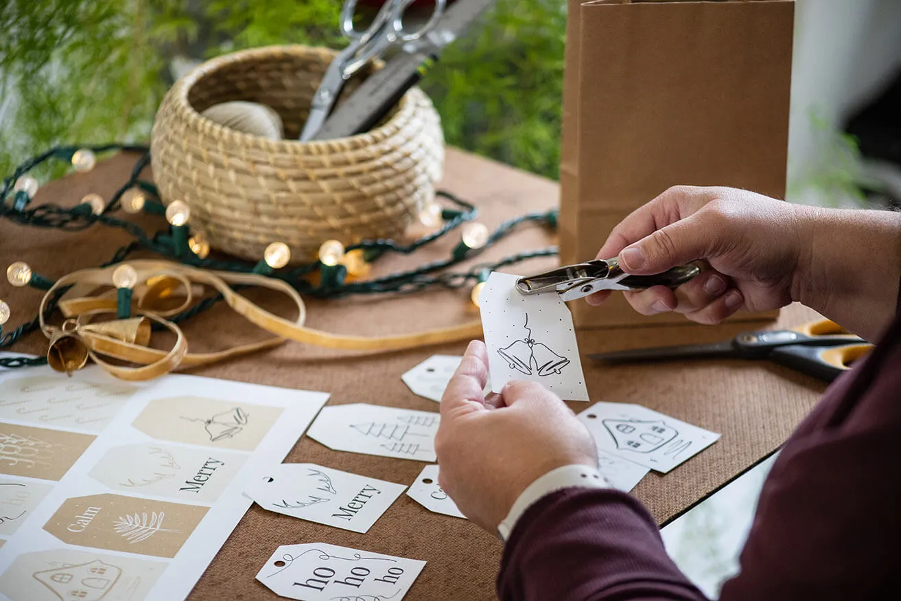 2022 Free Christmas Gift Tag Printables - myheartcreative A woman uses a handheld hole-puncher on a printable gift tag created by a boutique web design studio.
