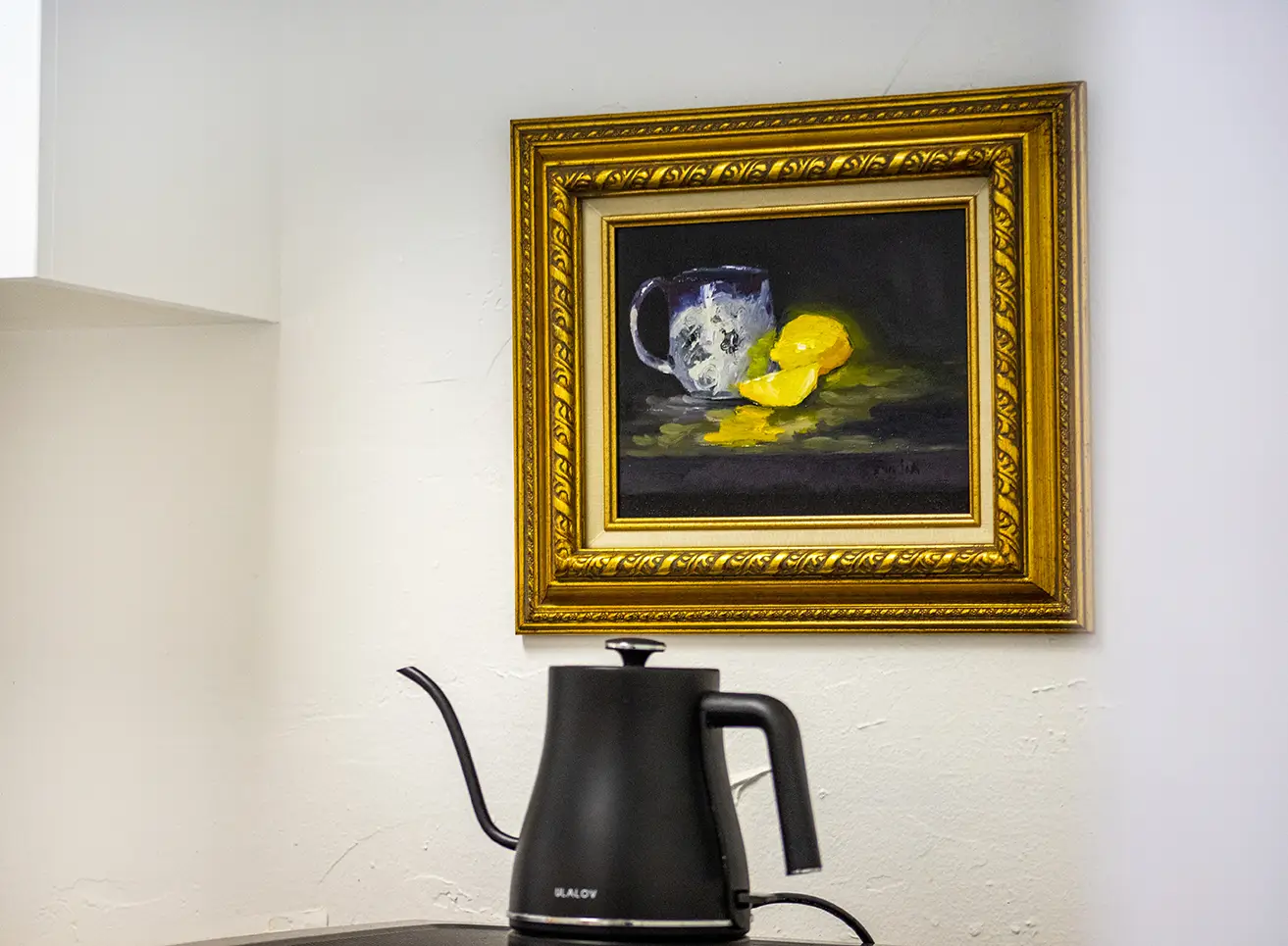 An acrylic painting of a coffee cup and lemons hangs on the wall in an ornate gold frame. A black electric tea kettle sits below it. 
