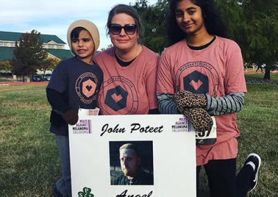 Amy and two children smile with a memorial sign after the Miles Against Melanoma race.