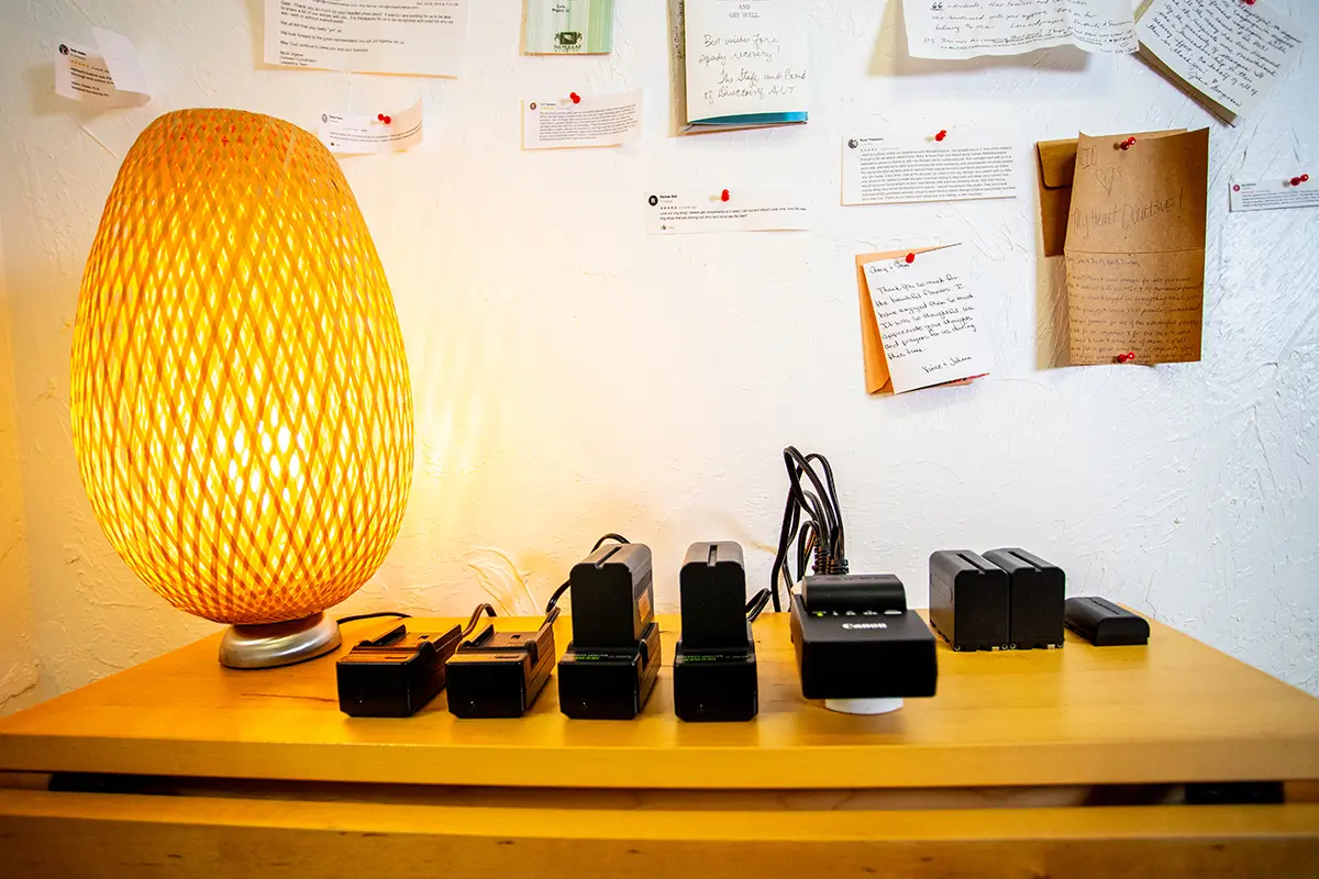 Differences Between Videography and Cinematography - myheartcreative Warmly lit studio desk with multiple camera battery chargers and Canon charging dock arranged beside a woven table lamp, with pinned client notes and handwritten letters displayed on the wall behind.