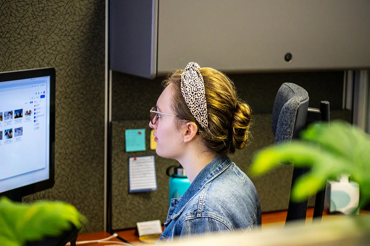 Is It Free to Own a Website? - myheartcreative Woman wearing glasses and a patterned headband working at a desktop computer in an office cubicle, viewing a website on the screen with sticky notes and a water bottle nearby.