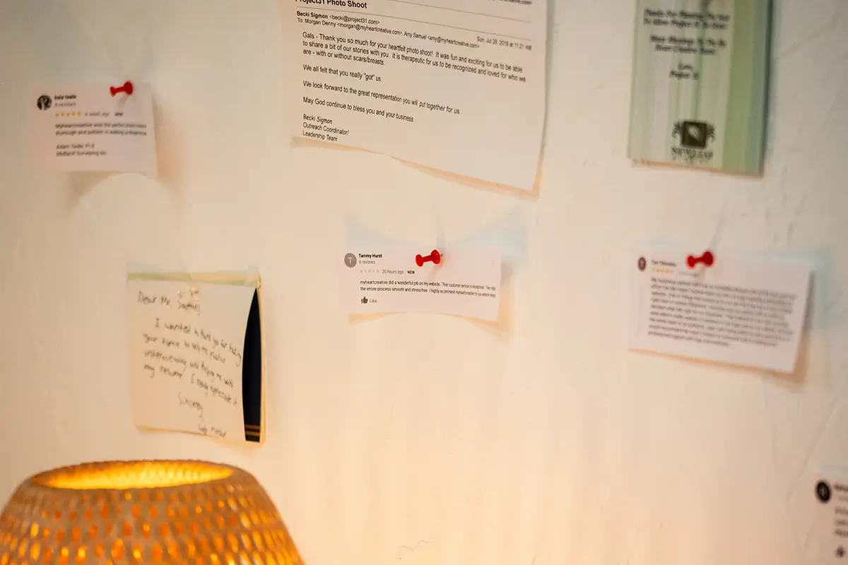 Client testimonials and printed reviews pinned to a white wall with red push pins above a warm woven table lamp in a creative studio space.