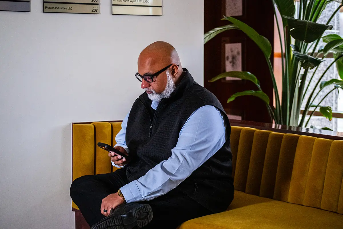 Man sitting in a modern office lobby on a mustard-yellow velvet bench, looking at his phone. He is wearing a black vest over a light blue dress shirt.