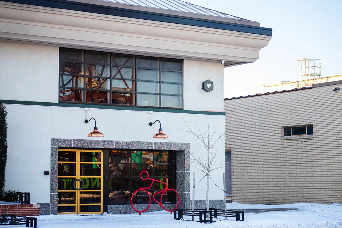 A modern white building in winter with copper gooseneck lights, a yellow-framed glass door, and a red stick-figure bicycle sculpture out front in the snow.