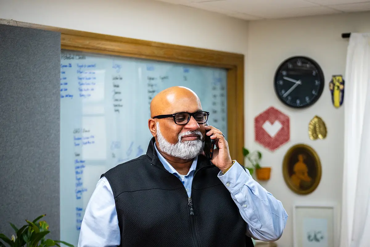 What Is Crawling in SEO? - myheartcreative A bald man with a grey beard and glasses smiles while talking on a phone. He wears a black vest over a light blue shirt, standing before a blurred office wall.