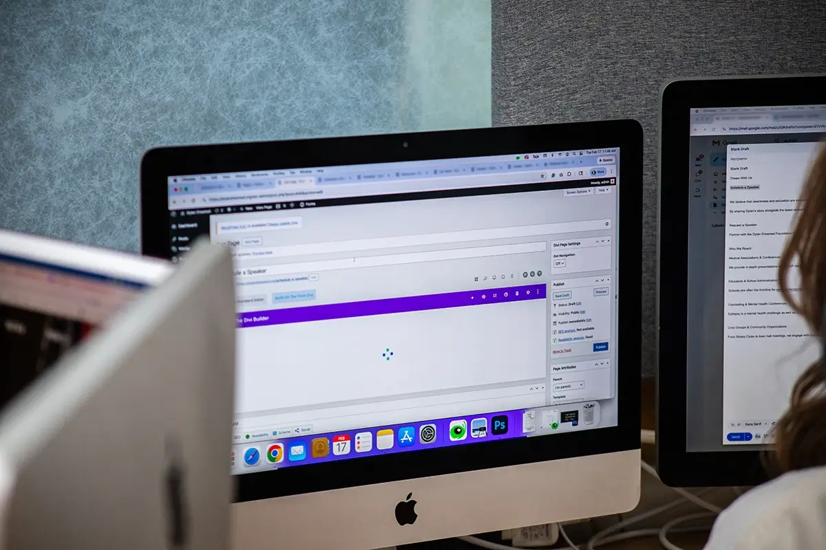 An iMac screen displaying a website builder interface with a purple toolbar, set in a modern office alongside other monitors and blurred equipment
